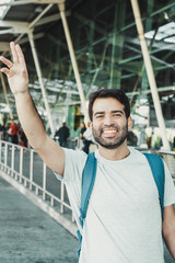 Fototapeta premium Smiling traveler waving hand near airport. Cheerful young man trying to stop car. Travel concept