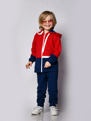 Happy smiling laughing blond kid boy in red and blue tracksuit sportwear and aviator sunglasses poses over white