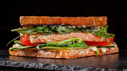 Vegetarian American Cuisine. Homemade toast sandwich with red fish salmon, arugula, cucumbers and tomatoes, white sauce. Sandwich on a black background. copy space text