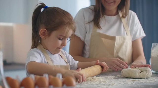 Medium shot of mother and daughter rolling dough in kitchen. Mom teaches daughter to cook dough. Girl learns to cook pastries. Kneading the dough together. Roll out the dough for baking.