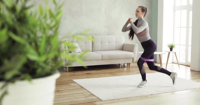 Sporty woman is training making squats and lunges at home in living room, side view. Sporty girl having workout. Training and making physical exercises with rope, fitness elastic band. Sport concept.