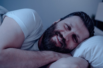 A lonely bearded dark-haired man with a very happy face lies on his side and looks at the camera at night on the bed. Close-up, date succeeded, happy, white bedding.