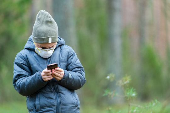 Kid With Mask In Their Mouths Due To Coronavirus Disease And Play Mobile Games On A Black Phone In A Forest