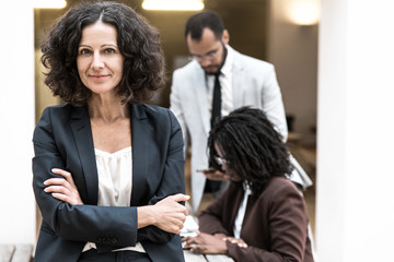 Friendly successful female professional posing with arms folded, looking at camera, smiling. Her multiethnic colleagues working in background. Female business leader concept