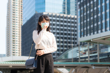 Fototapeta premium Young Asian businesswoman in shirt going to work feeling sick with cough wears protection mask prevent PM2.5 dust, smog, air pollution and COVID-19 with business office building in Bangkok, Thailand.