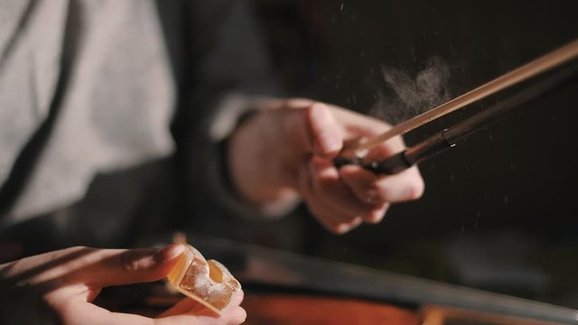 Young woman fiddler applying rosin to the violin bow hair to create friction between the bow and the strings, closeup shot, dust specks particles floating in air