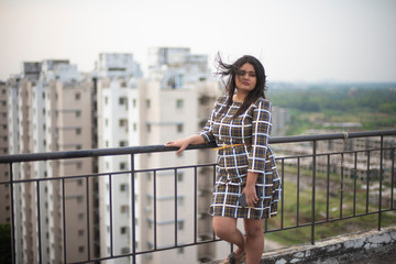 Fototapeta na wymiar An young brunette Indian Bengali brunette plus size woman in western dress standing on rooftop/balcony in urban background while her hair is blowing in wind during sunset. lifestyle and fashion.