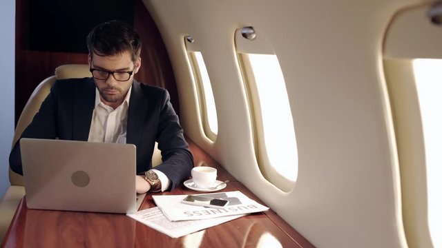 serious businessman using laptop near newspapers and coffee cup in plane
