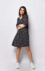 Fototapeta premium Brunette lady in black dress with floral print and white sneakers. Smiling, hugging herself, posing isolated on white. Full length
