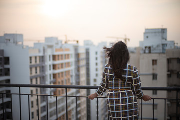 Plakat Back side of an young brunette Indian Bengali brunette plus size woman in western dress standing on rooftop in urban background while her hair is blowing in wind during sunset. lifestyle and fashion.