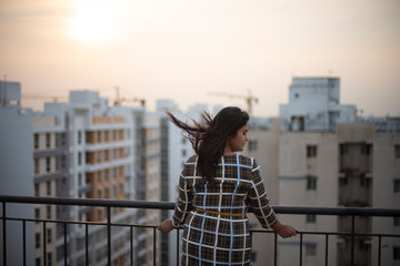 Back side of an young brunette Indian Bengali brunette plus size woman in western dress standing on rooftop in urban background while her hair is blowing in wind during sunset. lifestyle and fashion.
