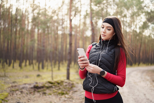 Caucasian girl is hiking at the weekend in a pine forest green and lush beautiful. She uses the running application on her smartphone. Healthy lifestyle concept. Modern lifestyle.