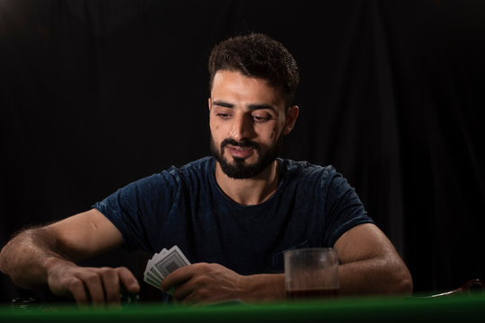 Portrait Of Young Brunette Indian Kashmiri Man In Casual Tee Shirt Playing Cards On A Casino Poker Table In Black Copy Space Studio Background. Lifestyle And Fashion.