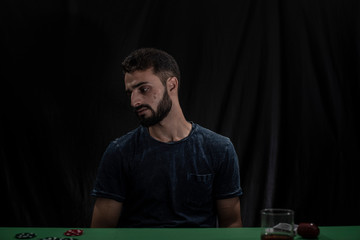 Portrait of young brunette Indian Kashmiri man in casual tee shirt playing cards on a casino poker table in black copy space studio background. lifestyle and fashion.