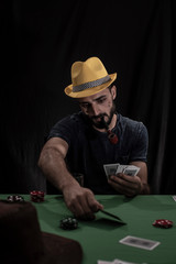 Portrait of young brunette Indian/European/Arabian/Kashmiri man in casual tee shirt and yellow hat playing cards on a casino poker table in black copy space studio background. lifestyle and fashion.