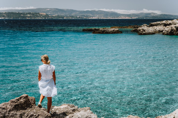 Adult female in white dress on summer vacation enjoying sea coast landscape of small beach with crystal clear blue water. Greece, Kefalonia