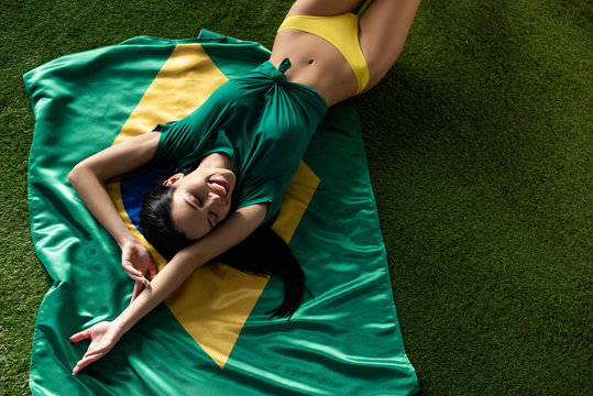 top view of smiling sexy girl lying on brazilian flag on green grass