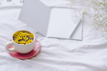 Сup with yellow flowers inside, on a white bed in the morning with notepad and pen. Spring background