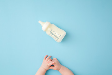 Infant hands and bottle of milk on light blue table background. Feeding time. Pastel color....