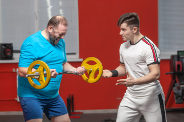 A fat man in the gym performs barbell lifting exercises at the limit of his powers under the guidance of a personal trainer. Overweight
