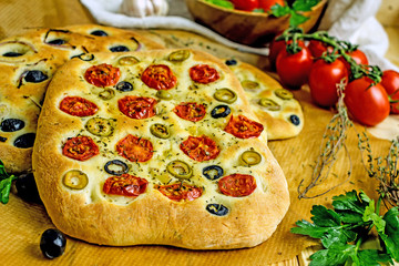 Italian focaccia with herbs, olives, garlic and tomatoes on a wooden table, next to fresh tomatoes, garlic, towel, sprigs of herbs. Homemade flat bread. Copy space, top view