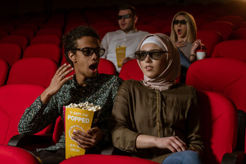 Obraz na płótnie Canvas African american muslim man with his wife sitting in movie theater, watching 3D movie, eating popcorn, smiling.