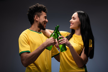 smiling multiethnic couple of football fans in yellow t-shirts clinking with bottles of beer on grey