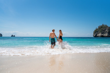 Fototapeta na wymiar Happy young couple enters the ocean with turquoise clear water. Paradise vacation of a guy and a girl in Bali. Romantic holidays on the island of Nusa Penida, Bali