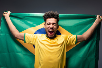 emotional african american football fan shouting and holding brazilian flag on grey