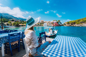 Fototapeta premium Holiday vacation. Woman in cafe enjoying time in Assos village in front of emerald bay of Mediterranean sea white boat and beautiful traditional houses of in Kefalonia, Greece