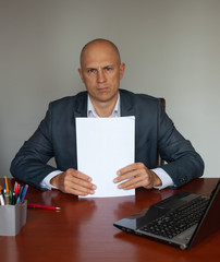 A serious man in a suit sits at a table with documents.