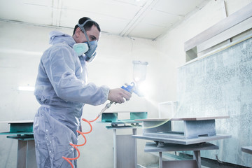 Process of production and manufacture of wooden furniture in factory. Worker man in overalls paints wood with paint spray - obrazy, fototapety, plakaty