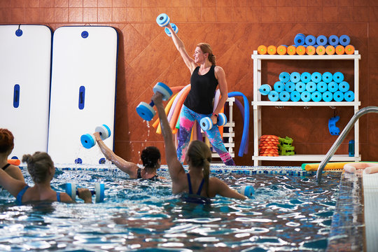 Young girls and middle-aged women do aqua aerobics with dumbbells in a swimming pool with a trainer, fitness exercises for weight loss in water