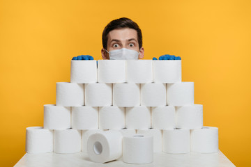 Scared man in a protective face mask is hiding behind toilet paper to protect himself from...