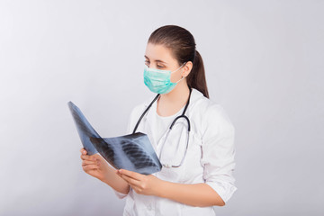 Attractive female doctor in a white coat, mask and with a stethoscope on his neck holds an x-ray in his hands.