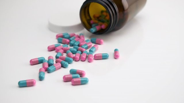 Close Up View of Teal Blue and Pink Colored Capsules and a Bottle Overturned on a White Background Moving Right to Left.