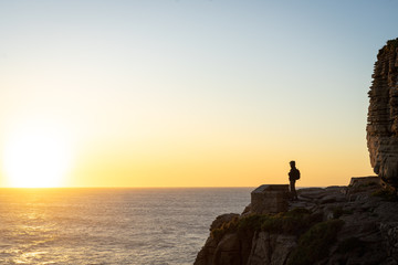 A person in the distance standing on the edge of a cliff looking at the ocean in the sunset. View from the side.