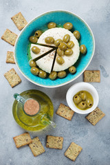 Green olives and soft cheese with black pepper, olive oil and crackers. Flatlay on a light-blue stone surface, vertical shot