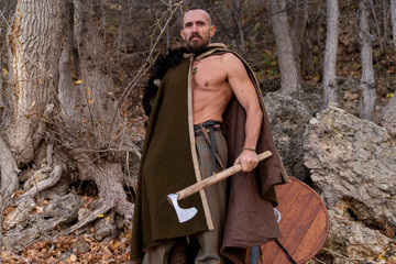 Naklejka premium Viking dressed in a cape and skin armed with an ax and a sword stands on the background of the forest