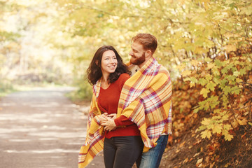 Beautiful couple man woman in love. Boyfriend and girlfriend wrapped in yellow blanket plaid hugging in a park on autumn fall day. Tenderness and happiness. Authentic real people.