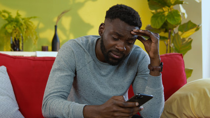 Close-up afro-american man messaging on smartphone sitting on sofa. Upset sick man feeling sudden pain suffering headache covering massaging face.