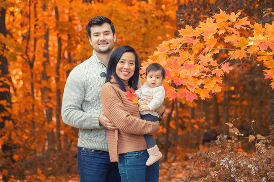 Asian Chinese Mother And Caucasian Father Dad With Baby Girl In An Autumn Fall Park. Family Together Outdoor In Nature. Seasonal Holiday And Authentic Lifestyle.