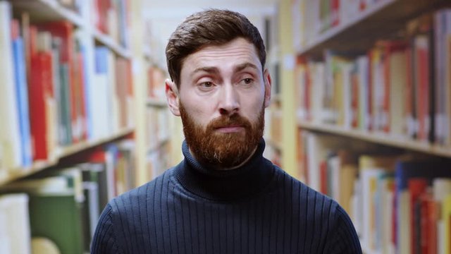 Doubtful young bearded man in blue jamper showing confused face shrugging his shoudlers to express misunderstanding working at a library.