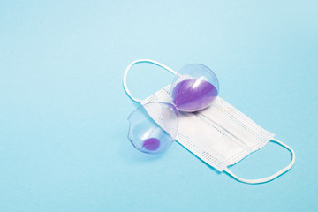 broken hourglass with purple sand and a medical protective white mask on a blue background copy space, deadly disease