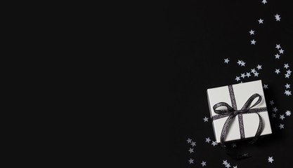 White gift box with silver ribbon and star shaped confetti on black background. Top view, copy space.