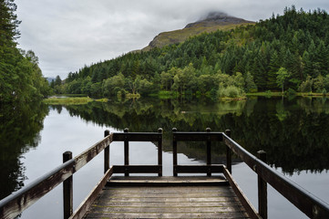Landscape, reflections and a wooden walkway upon Glencoe Lochan on a cloudy day, in the Scottish Highlands, Scotland. 