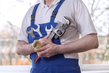Mężczyzna hydraulik w białej koszulce polo i niebieskich spodniach ogrodniczkach trzyma w kieszeni na piersi klucze płaskie. W dłoniach trzyma pakuły i klucz nastawny, żabkę,