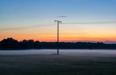 Fototapeta na wymiar Electricity Pylon with Power Lines in the Middle of a Field against the Background of Evening Fog and Haze at Sunset