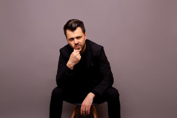 Pensive elegant man in black business suit sitting in studio with palms touching his beard while resting on chair on grey background. Copy space.