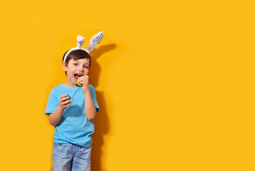 Naklejka premium Portrait of a smiling adorable little boy with bunny ears and holding colorful easter eggs isolated orange background